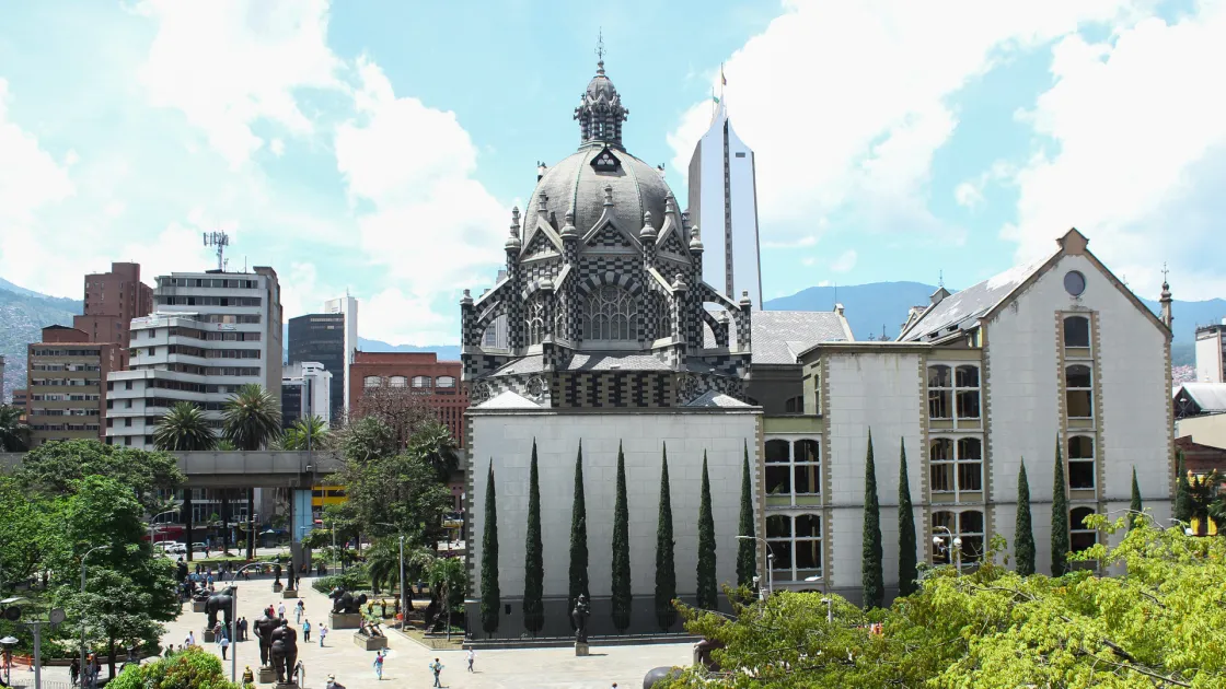 La imagen destaca la impresionante arquitectura neogótica flamenca del Palacio de la Cultura, con su icónico patrón de cuadros blancos y negros ("fachada ajedrezada") y su gran cúpula central. En primer plano, se observa la Plaza Botero, un museo al aire libre que alberga las famosas esculturas de bronce de Fernando Botero, rodeadas de transeúntes y palmeras delgadas.