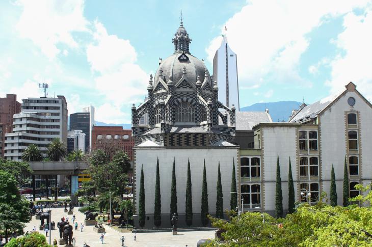 La imagen destaca la impresionante arquitectura neogótica flamenca del Palacio de la Cultura, con su icónico patrón de cuadros blancos y negros ("fachada ajedrezada") y su gran cúpula central. En primer plano, se observa la Plaza Botero, un museo al aire libre que alberga las famosas esculturas de bronce de Fernando Botero, rodeadas de transeúntes y palmeras delgadas.