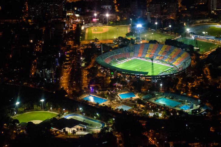 La imagen muestra el complejo deportivo iluminado bajo la oscuridad de la noche. En el centro destaca el Estadio Atanasio Girardot, con su gramado verde brillante y gradas coloridas. A su alrededor se aprecian otras instalaciones, como diamantes de béisbol, canchas auxiliares y un complejo acuático con varias piscinas iluminadas de azul. Las luces de la ciudad y el tráfico en las avenidas circundantes completan la escena urbana.
