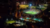 La imagen muestra el complejo deportivo iluminado bajo la oscuridad de la noche. En el centro destaca el Estadio Atanasio Girardot, con su gramado verde brillante y gradas coloridas. A su alrededor se aprecian otras instalaciones, como diamantes de béisbol, canchas auxiliares y un complejo acuático con varias piscinas iluminadas de azul. Las luces de la ciudad y el tráfico en las avenidas circundantes completan la escena urbana.